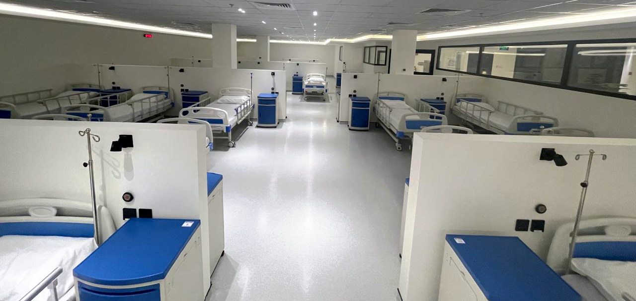 Wide view of the NAMA Bio-Center participant ward, with rows of blue-and-white clinical beds and bedside cabinets in partitioned bays under bright ceiling lighting.