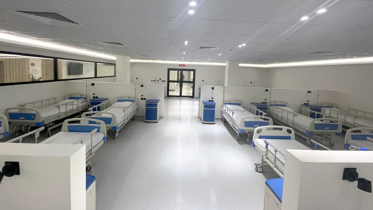 The participant ward at NAMA Bio-Center seen straight down its central aisle, with partitioned bays of blue-and-white clinical beds and bedside cabinets running away on both sides.