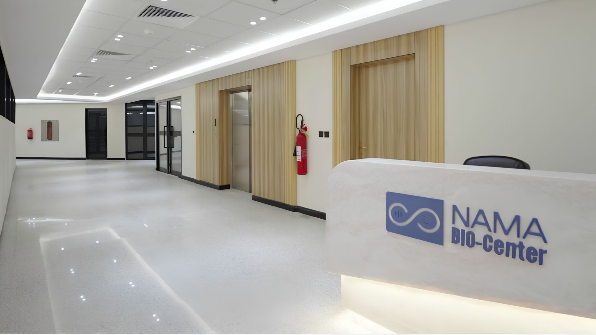 The NAMA Bio-Center reception lobby: a pale stone desk carrying the blue NAMA BIO-Center lockup, facing a bright polished corridor with timber-panelled lift doors.