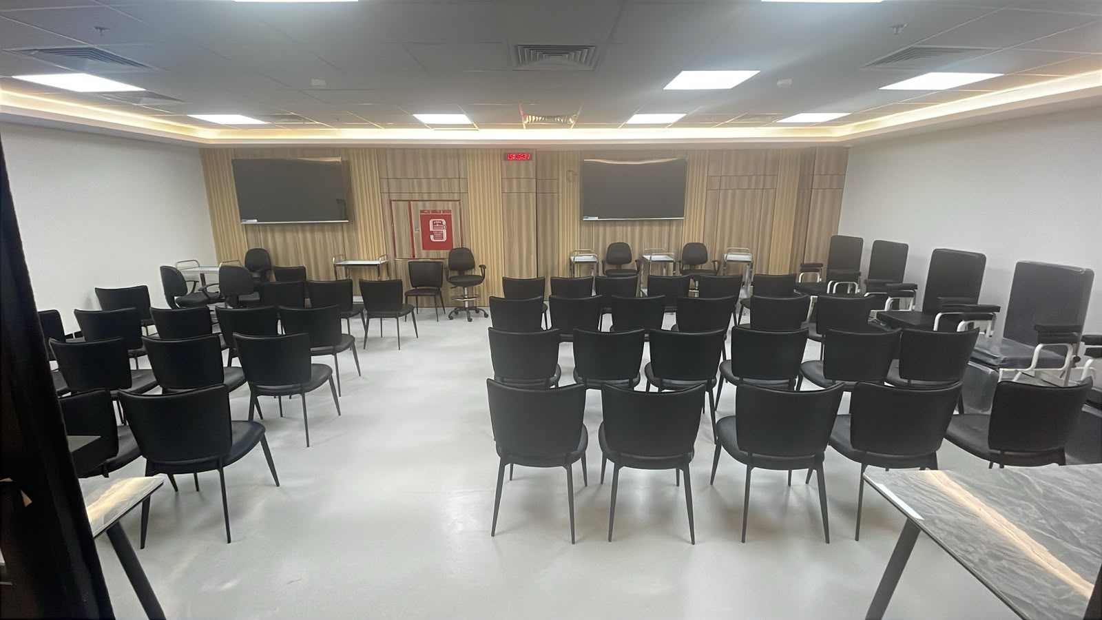 The NAMA Bio-Center training room from the back of the seating, with rows of black chairs and side writing tables facing two wall-mounted screens on the wood-panelled front wall.